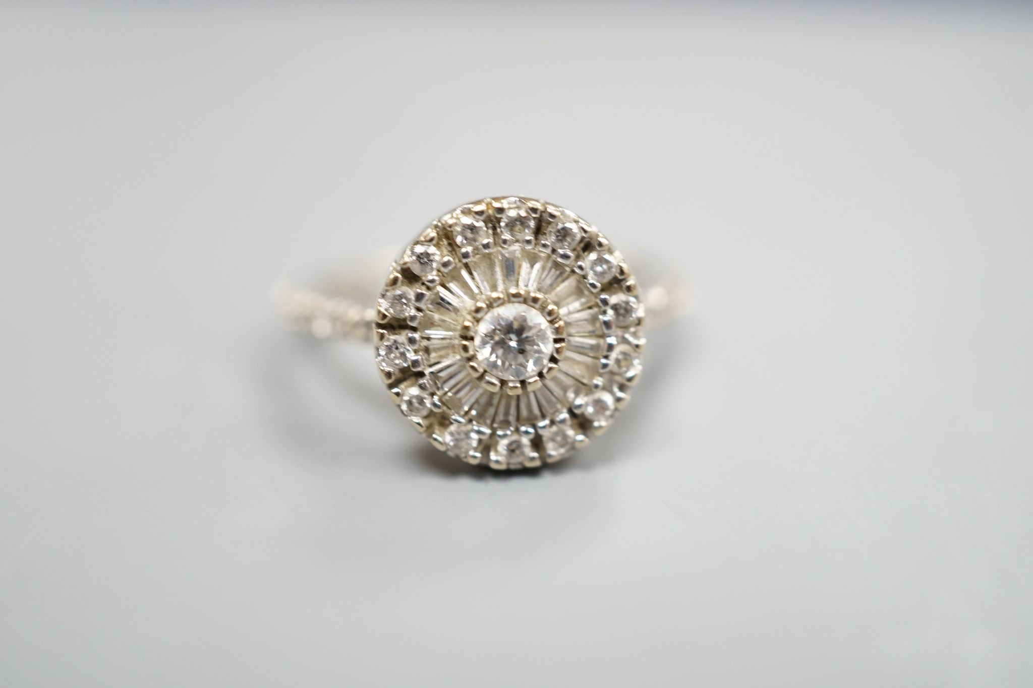 A modern white metal, round and trapeze cut diamond set target cluster ring, with diamond set shoulders, size N, gross weight 4.2 grams.
